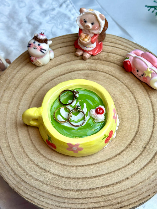 Yellow buttercup matcha ring holder cup with green beverage and two rings on a woven mat with small decorative figures.