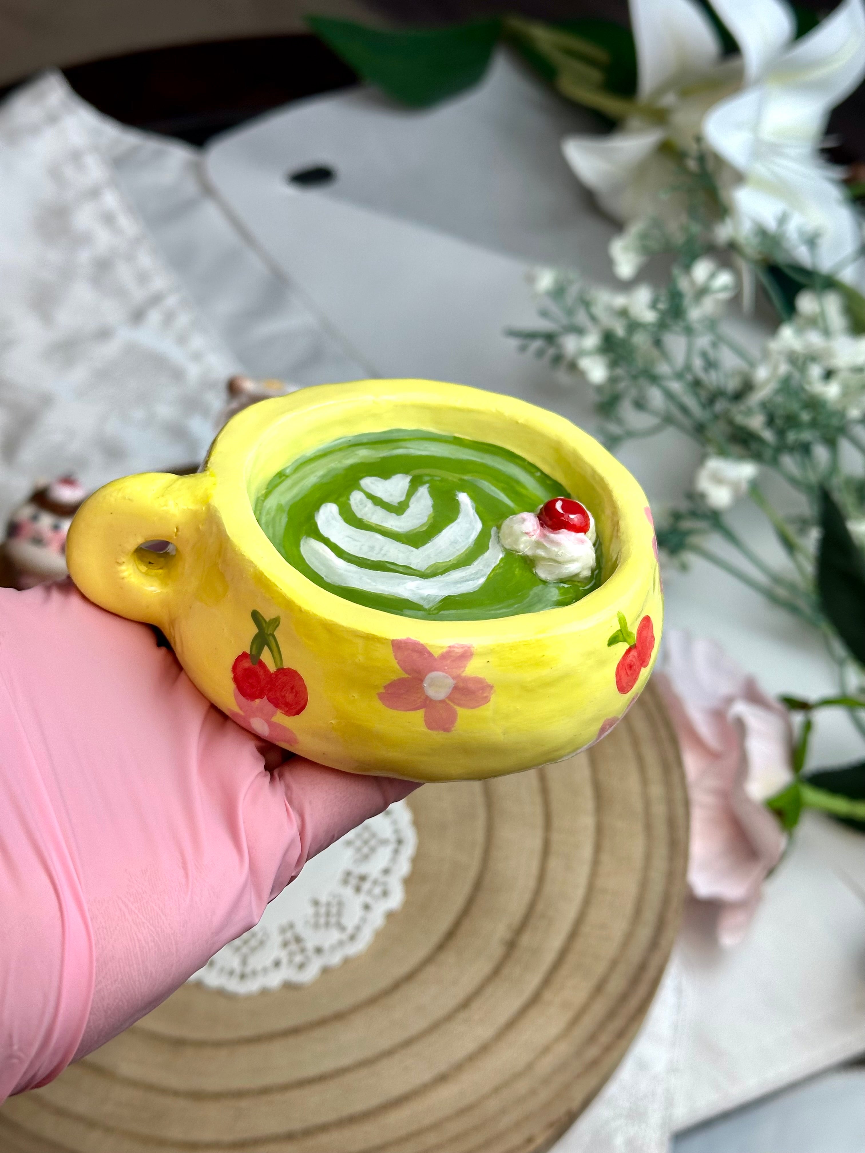 Yellow buttercup matcha ring holder cup with green beverage and cherry decoration, held by a hand.